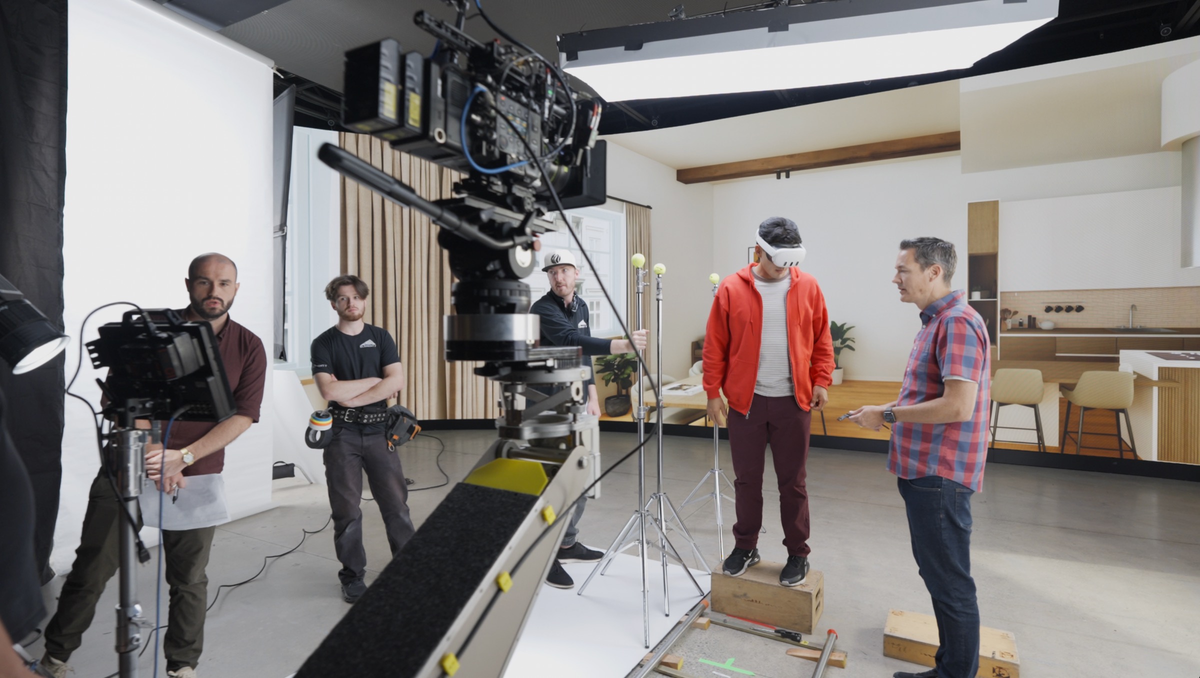 Behind-the-scenes on-set — talent wearing white XR headset on LED volume stage with modern living room interior on LED wall, director giving direction, cinema camera on robotic motion control arm with VFX tracking markers